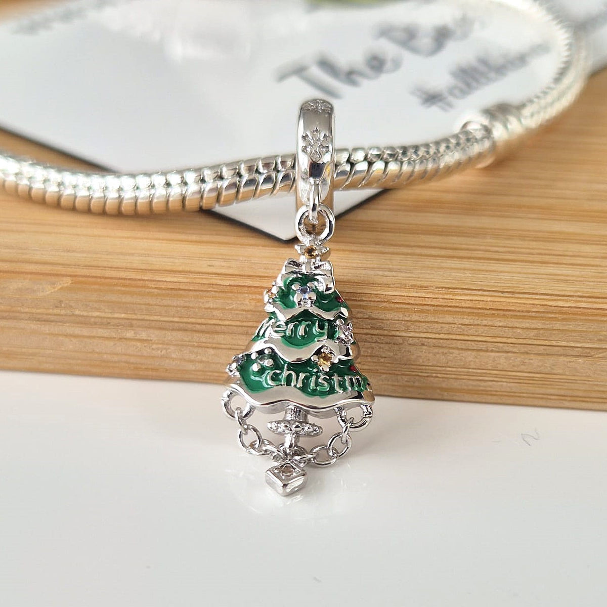 A silver Christmas tree charm with green enamel and 'Merry Christmas' lettering, shown hanging from a silver snake chain bracelet on a wooden surface.