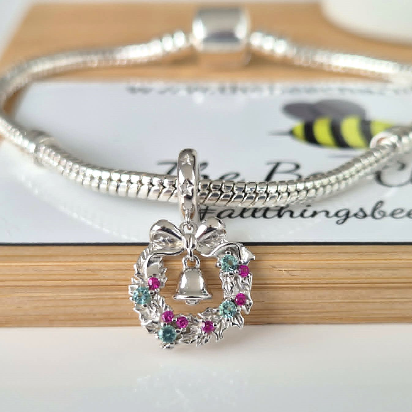 A silver snake chain bracelet with a wreath pendant featuring a silver bell and pink and blue gemstones, displayed on a wooden surface with a branded card underneath.