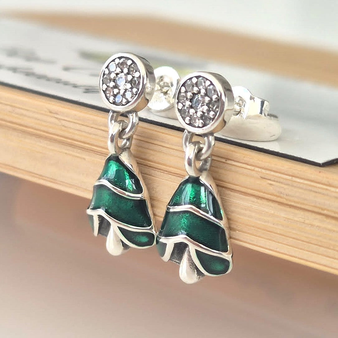 A pair of silver-toned drop earrings with round crystal-studded tops and dangling tree-shaped charms filled with glossy green enamel, resting on a light wood surface.