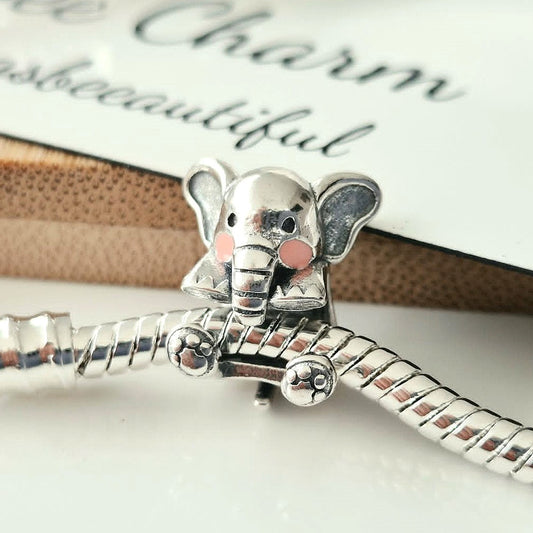 A silver elephant charm with pink cheek details on a silver snake chain, resting on a white surface with a card in the background.